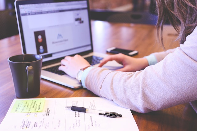 a person working with a laptop, with a notebook, pen, and cup on the table